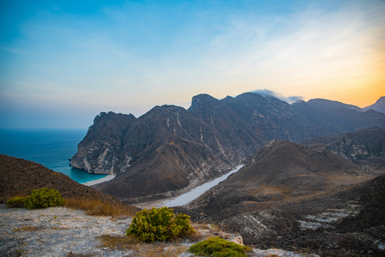 Dramatic golden-hour view over rugged Oman mountains plunging to a turquoise cove and fjord-like inlet at sunset